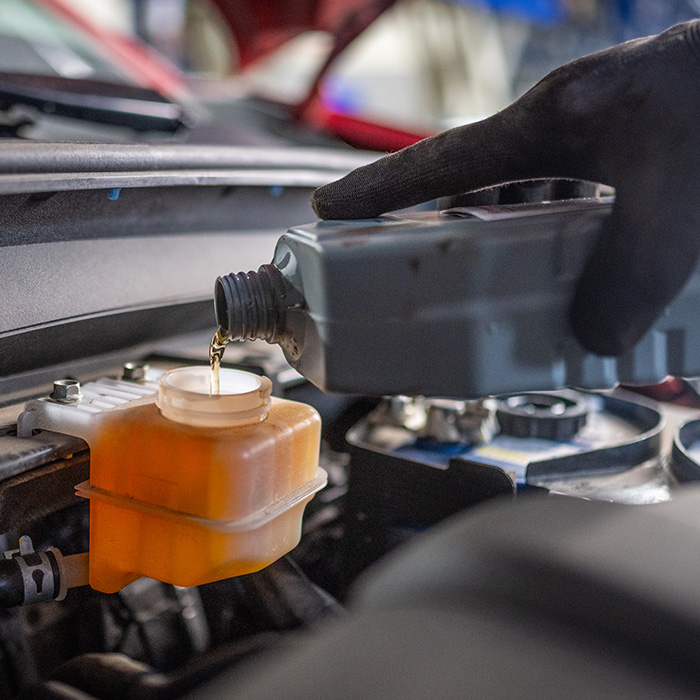 A mechanic is refilling brake fluid in a car engine bay, wearing protective gloves